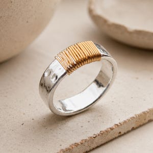 May include: A silver-toned ring featuring a hammered texture on the band and a section wrapped with thin gold-toned wire. The ring has a polished finish and a circular shape, showcasing a contrast between the metallic silver band and the gold wire.