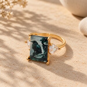May include: A gold-toned ring featuring a rectangular center stone with a dark green and black graphic print of a raven perched on a rock. The ring is accented by two clear, triangular-cut stones on either side of the central rectangular setting.