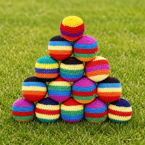 Puede incluir: Una pirámide de pelotas de malabares coloridas, hechas a mano con ganchillo. Cada pelota presenta rayas horizontales en colores del arcoíris, incluyendo rojo, naranja, amarillo, verde, azul y morado. Las pelotas están dispuestas sobre hierba verde.