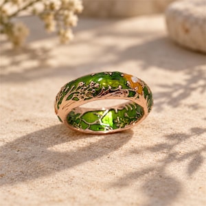 May include: A rose gold band ring featuring an intricate green enamel inlay depicting a forest landscape with tree branches and foliage. A small yellow flower with a white center is visible on the top surface of the metal ring.