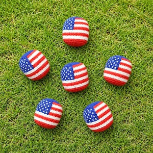 May include: Six red, white, and blue crocheted balls, each featuring the American flag design. The balls are scattered on a bed of green grass, showcasing the patriotic theme. The balls are a fun, decorative item.