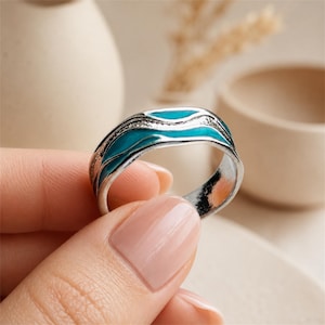 May include: A silver-toned metal ring featuring a wavy, organic design with inlaid bright turquoise enamel accents. The band has a textured, slightly irregular surface that follows the flowing lines of the blue enamel inlay throughout the ring.