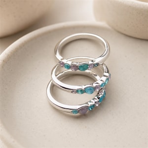 May include: A stack of three silver rings, each featuring a row of small, round, bezel-set gemstones in alternating shades of light blue, turquoise, and pale lavender. The bands are smooth and polished, creating a cohesive set of jewelry.
