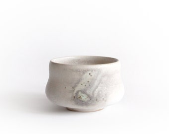 May include: A white ceramic teacup with a speckled glaze. The cup has a rounded shape and a slightly flared lip.