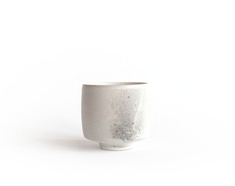 May include: A white ceramic cup with a slightly flared rim and a wide base. The cup has a subtle, mottled glaze that gives it a unique, handcrafted look.