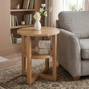 May include: A light oak side table with a circular top and two shelves. A white vase with flowers is on top, and two decorative boxes are on the lower shelf. The table has a cross-shaped base. The table is next to a grey sofa.