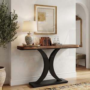 May include: A console table with a rectangular brown wood top and a black metal base featuring two curved legs. On the table sits a white lamp with a patterned base, a gold tray, five upright books, and two clear glass decanters.