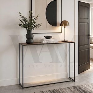 May include: A rectangular console table with a dark wood top and a thin black metal frame. On the table sits a dark grey vase with green eucalyptus branches, a small black bowl, and two stacked books labelled Chanel and Tom Ford 002.