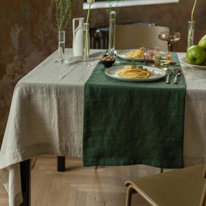 Könnte beinhalten: Ein rechteckiger Tischläufer aus dunkelgrünem Leinen, der über einer hellbeigen Leinentischdecke liegt. Der Tisch ist mit weißen Keramiktellern mit Pasta, klaren Glasvasen, einer kleinen schwarzen Schale mit Nüssen und Besteck gedeckt.