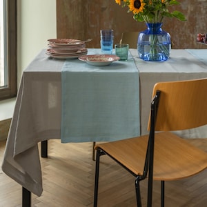 Puede incluir: Un mantel de lino gris claro con un camino de mesa de lino azul claro. Sobre la mesa hay una pila de platos blancos con motivos rojos, un jarrón de cristal azul con girasoles y dos vasos pequeños de cristal azul y verde sobre una mesa.