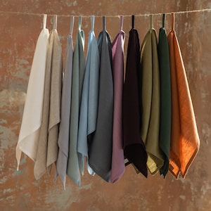 May include: A collection of eleven rectangular kitchen towels hanging from a horizontal string. The towels are made of a textured fabric and come in a variety of colors including white, beige, light blue, grey, purple, dark brown, olive, and orange.