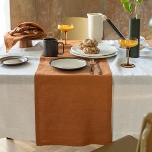 May include: A rectangular rust-colored linen table runner centered on a white tablecloth. The setting includes a black mug, a white plate with seeded rolls, a fork, a knife, two amber-colored stemmed glasses, and a white pitcher.