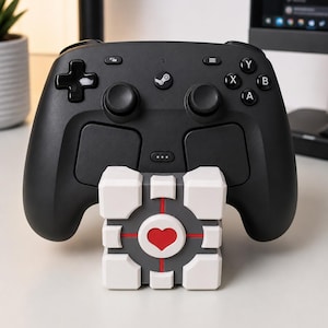 May include: A black Steam controller with a white and gray companion cube. The controller has a D-pad, two joysticks, and action buttons. The companion cube has a red heart in the center and is a popular gaming reference.