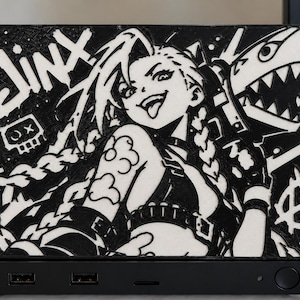 Puede incluir: Un panel decorativo rectangular en blanco y negro que presenta una ilustración gráfica del personaje Jinx con cabello largo trenzado, un arma con forma de tiburón y el texto JINX. El panel está montado sobre una base negra con dos puertos USB.