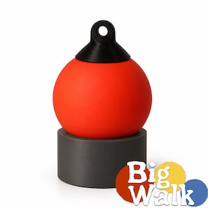 Op de afbeelding: Een bolvormig rood object met een zwarte bovenkant voorzien van een cirkelvormige ophanglus, rustend op een cilindrische donkergrijze basis. Het object heeft een gladde matte afwerking. Rechtsonder staat een logo met de tekst Big Walk.