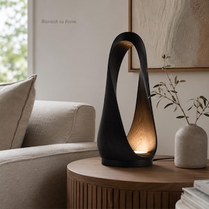 May include: A sculptural table lamp with a dark, curved wooden frame that forms an elongated, open loop. The interior of the frame features a natural wood grain finish, illuminated by a light source at the base. The text "Warmth in Form" is visible.