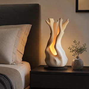 May include: A table lamp shaped like a hollowed-out tree trunk with a light-coloured wood grain finish. The lamp emits a warm, soft glow from within the central opening and sits on a circular dark base, casting light upward through the branches.