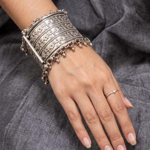 Könnte beinhalten: Ein breites, silberfarbenes Manschettenarmband mit detaillierten Blumenprägungen auf mehreren horizontalen Bändern, verziert mit kleinen, hängenden kugelförmigen Anhängern an den Rändern, kombiniert mit einem dünnen, schlichten silberfarbenen Ring.