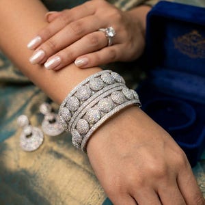 May include: A set of three silver-toned bangles featuring circular, pavé-set crystal embellishments. The jewelry set includes matching circular drop earrings and a solitaire ring with a clear stone, all displayed with a dark blue velvet box.