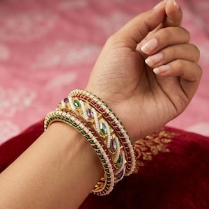 Puede incluir: Un conjunto de tres brazaletes dorados con intrincados abalorios en rojo, verde y blanco. El brazalete central presenta piedras de talla marquesa en tonos morados y verdes, mientras que los brazaletes exteriores están bordeados por filas de cuentas.