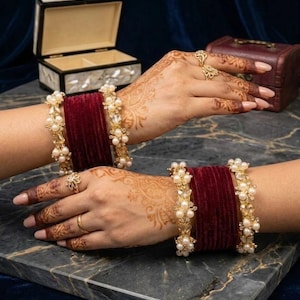 Pode incluir: Um conjunto de pulseiras de veludo vermelho escuro acentuadas com metal dourado e pérolas artificiais brancas. O conjunto de joias inclui anéis a combinar com metal dourado e pérolas artificiais brancas, desenhados como acessórios coordenados.