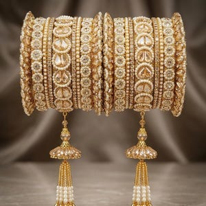 May include: A set of gold-toned bangles featuring intricate patterns of clear teardrop-shaped stones and small white pearls. The bangles are arranged in a stack and include two dangling bell-shaped charms adorned with crystals and pearl beads.
