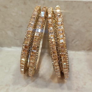 Può includere: Un set di quattro bracciali rigidi color oro con una fila centrale di cristalli quadrati color ambra, affiancati da due file più sottili di piccole pietre trasparenti incastonate a pavé. I bracciali circolari presentano una finitura metallica.