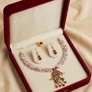 May include: A jewellery set featuring a multi-strand necklace with white and maroon beads, a gold-toned pendant depicting a flute, matching drop earrings with maroon beads, and a square ring with a maroon stone, all presented in a velvet box.