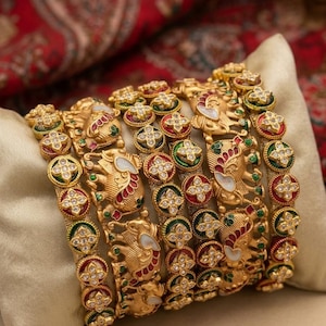 May include: A set of gold-toned metal bangles featuring embossed elephant motifs with white, red, and green enamel accents. The bangles are decorated with small, clear, sparkling stones arranged in floral-shaped settings along the edges.