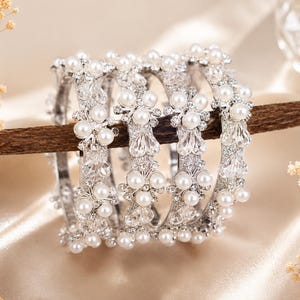 May include: A set of four silver-toned bangles featuring a repeating pattern of white faux pearls and clear faceted crystals. Each bangle is constructed with a metallic base and adorned with clusters of round pearls and sparkling crystal accents.