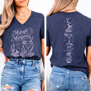 May include: A dark blue heathered V-neck t-shirt featuring a light purple graphic print. The front displays the text Midnight Margaritas with a candle, frog, and cocktail glass. The back shows a vertical column of icons including a moon, frog, cocktail, coffee pot, mushroom, and citrus slice.