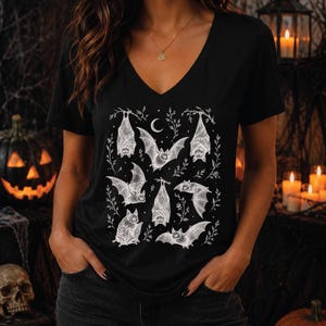 May include: A black V-neck t-shirt featuring a white graphic print of multiple bats in various poses, including hanging and flying, interspersed with small botanical sprigs and a crescent moon. The design is centered on the front of the shirt.