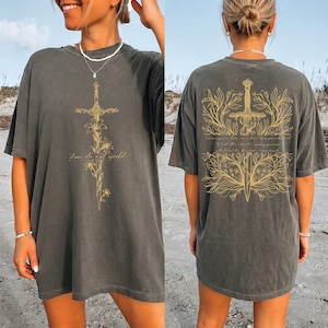 May include: Dark gray t-shirt featuring a gold sword design with floral accents on the front and back. The front of the shirt has the text "You do not yield." The back of the shirt has a sword design with text. The shirt is a relaxed fit.