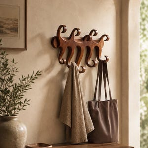 Könnte beinhalten: Eine hölzerne Wandgarderobe mit einer Reihe geschwungener, schnörkelartiger Haken. Die Garderobe ist aus mittelbraunem Holz mit glatter Oberfläche gefertigt. Ein beiges Handtuch und eine braune Ledertasche hängen an den Haken der Garderobe.