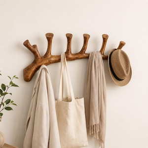 May include: A wall-mounted coat rack crafted from a single piece of natural brown wood, featuring multiple upward-curving branches as hooks. A beige jacket, a canvas tote bag, a light brown scarf, and a straw hat are hanging from the hooks.
