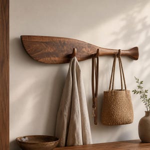 Könnte beinhalten: Eine Wandgarderobe in Form eines hölzernen Bootsruders mit dunkelbrauner Holzmaserung. Die Garderobe verfügt über drei geschwungene Haken, an denen ein beiges Leinenkleidungsstück, ein brauner Lederriemen und eine geflochtene Strohtasche hängen.