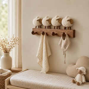 Könnte beinhalten: Eine hölzerne Wandgarderobe mit vier abgerundeten Haken und vier dekorativen cremefarbenen Schaf-Figuren mit braunen Gesichtern und Beinen an der Oberkante. Ein helles Musselintuch und ein kleines Lätzchen hängen an den Haken.