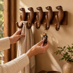 Könnte beinhalten: Eine Wandgarderobe aus Holz mit fünf geschnitzten Erdmännchenfiguren, deren geschwungene Schwänze als Haken dienen. Die Leiste besteht aus dunkelbraunem Holz mit strukturierter Oberfläche. Ein Haken hält ein beiges Tuch, ein anderer einen Schlüsselbund.