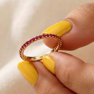 Puede incluir: Un anillo de banda fina de oro con una hilera continua de pequeñas piedras preciosas de color rojo vibrante engastadas a lo largo de la circunferencia exterior. El interior tiene grabado el texto 14K, indicando la pureza del oro.