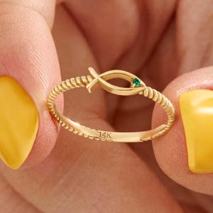 Könnte beinhalten: Ein goldener Ring mit einem gedrehten, seilartigen Band und einem zentralen fischförmigen Motiv. Das Fischdesign enthält einen kleinen, runden grünen Edelstein in der Nähe des Schwanzes. Die Innenseite des Rings ist deutlich mit 14K geprägt.