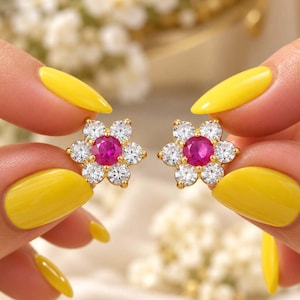 Könnte beinhalten: Ein Paar goldene Ohrstecker in Blumenform, jeweils mit einem zentralen, rund geschliffenen, leuchtend pinkfarbenen Edelstein, der von sechs klaren, funkelnden weißen, rund geschliffenen Steinen umgeben ist.
