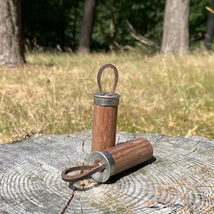 Op de afbeelding: Twee cilindervormige houten sleutelhangers met zilverkleurige metalen doppen en bruine leren lussen. Het hout heeft een zichtbare nerf en de metalen doppen zijn voorzien van gegraveerde tekst. Eén sleutelhanger staat rechtop, de andere ligt.