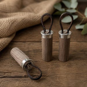 Pode incluir: Três objetos cilíndricos de madeira castanho-escura com tampas de metal prateado no topo. Cada tampa possui uma pequena alça de couro castanho-escuro fixada ao centro. Dois objetos estão na vertical, enquanto um está na horizontal.