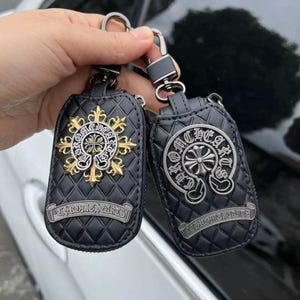 May include: Two black leather keychains with silver and gold cross designs. The keychains have a quilted pattern and the text "CHROME HEARTS" on a banner. Each keychain has a metal clasp.