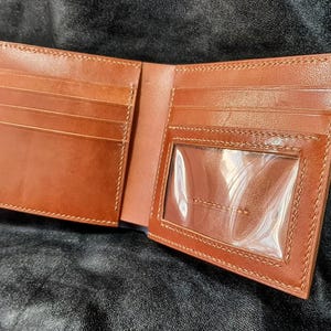 May include: Open, brown leather wallet with multiple card slots and a clear ID window. The wallet is made of smooth, stitched leather and is displayed open, showcasing its interior design.