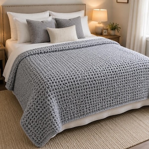 May include: A chunky knit throw blanket in a solid light gray color with a visible grid-like texture. The blanket is draped over a bed featuring white sheets, two large white pillows, two gray square pillows, and one small rectangular cream pillow.