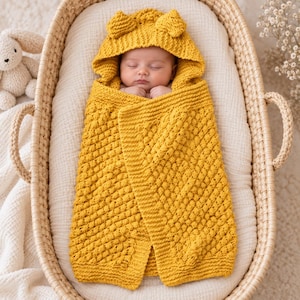 May include: A mustard yellow knitted baby swaddle blanket featuring a textured bobble stitch pattern and an attached hood with two small rounded ears on top. The blanket is wrapped securely around a sleeping infant, providing full coverage.