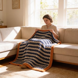 May include: A rectangular crochet blanket featuring horizontal stripes in navy blue and cream, finished with a thick, textured border in a vibrant burnt orange yarn. The blanket displays a consistent, repeating stitch pattern throughout the design.
