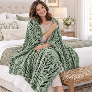 May include: A sage green crochet throw blanket featuring a repeating chevron stitch pattern with an openwork design. The blanket is draped over a person wearing a light beige knit top, showcasing the textured yarn and intricate stitch work.