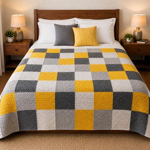 Puede incluir: Una manta de ganchillo con un patrón de cuadros repetitivo de bloques cuadrados en amarillo mostaza, gris carbón oscuro y gris claro. La manta está extendida sobre una cama, mostrando una apariencia texturizada y tejida a mano con cuadrados uniformes.